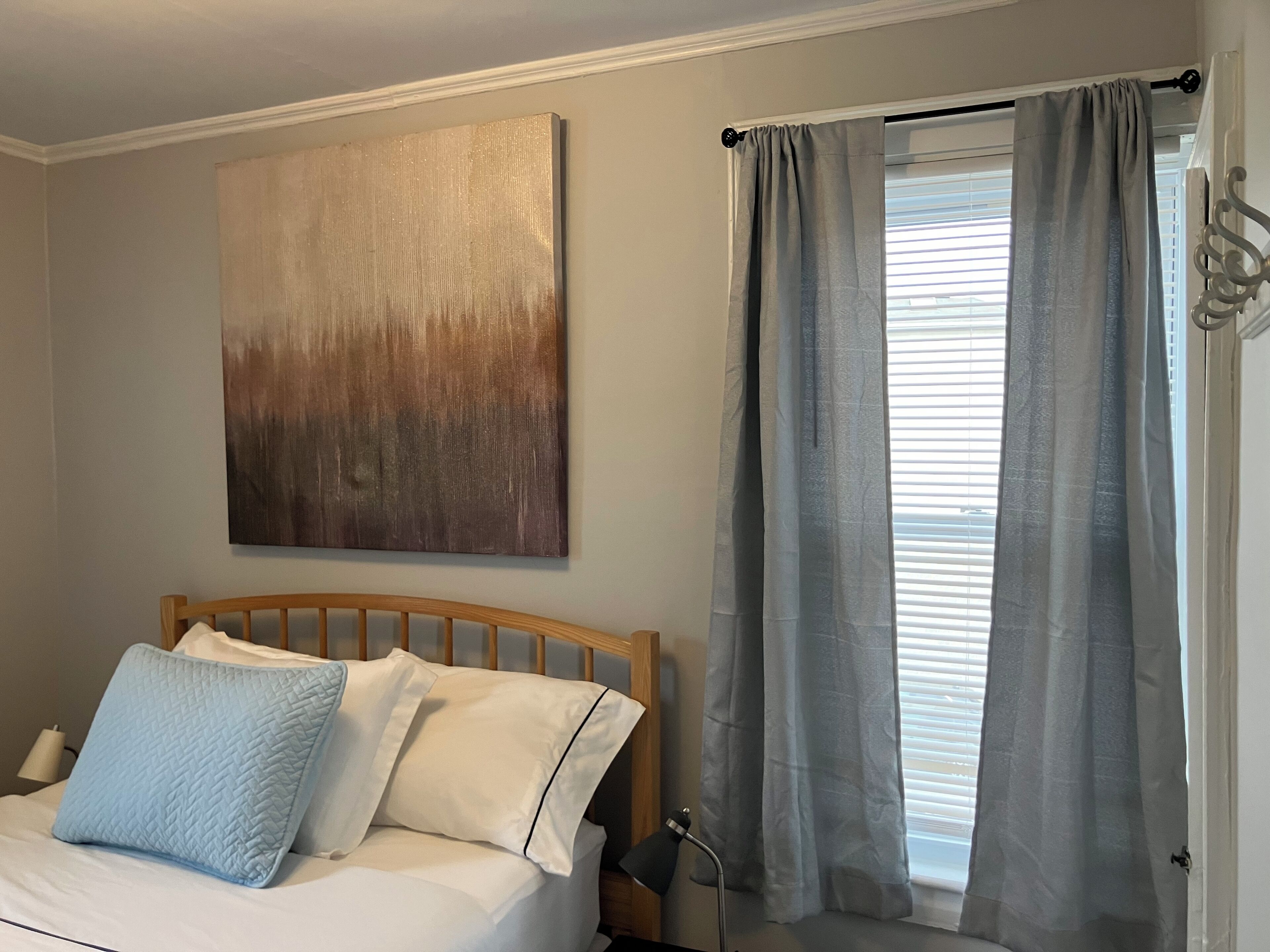 Een slaapkamer met een bed, een bedlamp, een raam met gordijnen en een aan de muur hangend abstract schilderij.