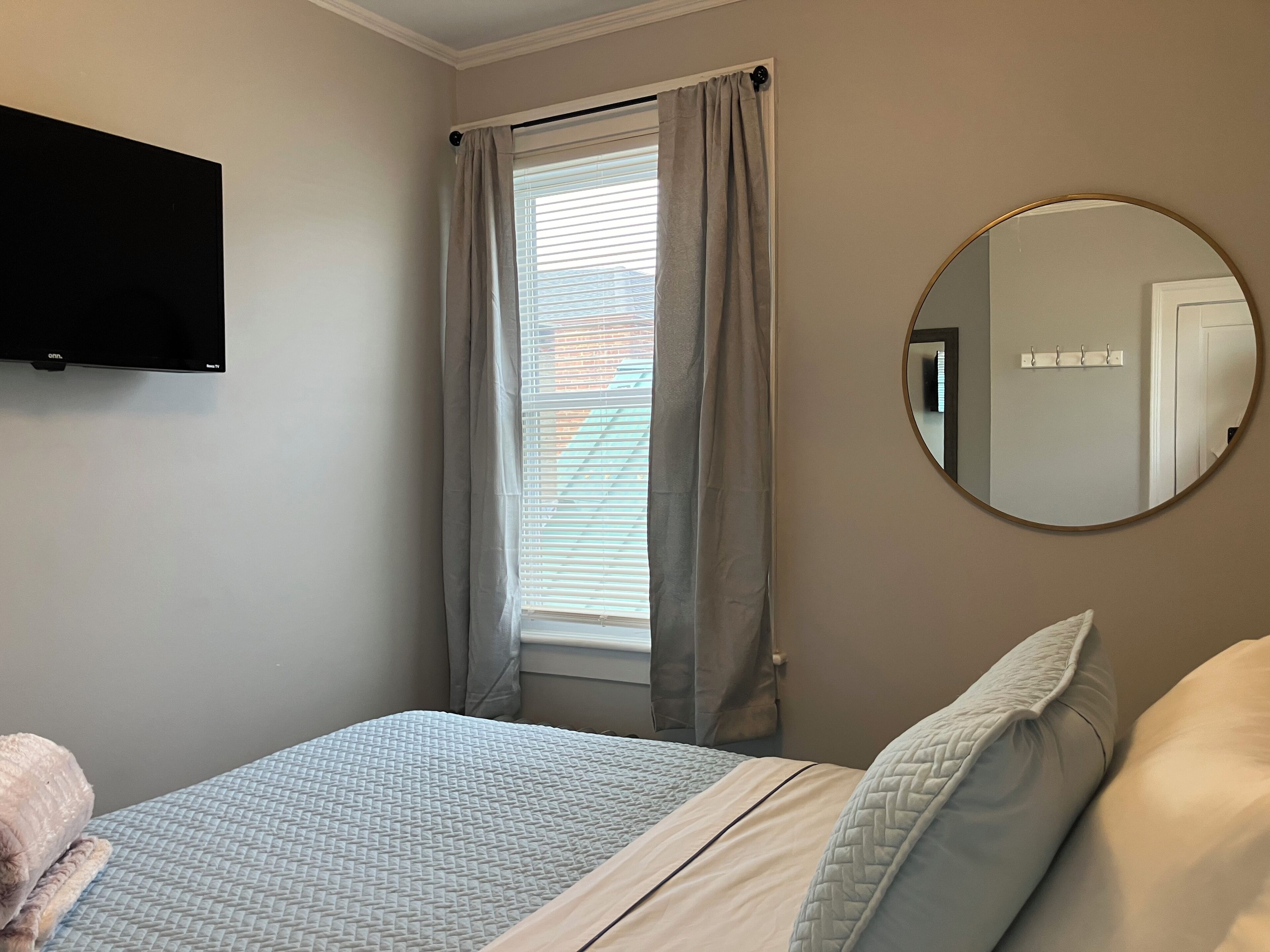 A bedroom with a bed, a round mirror, a window with curtains, and a wall-mounted TV.