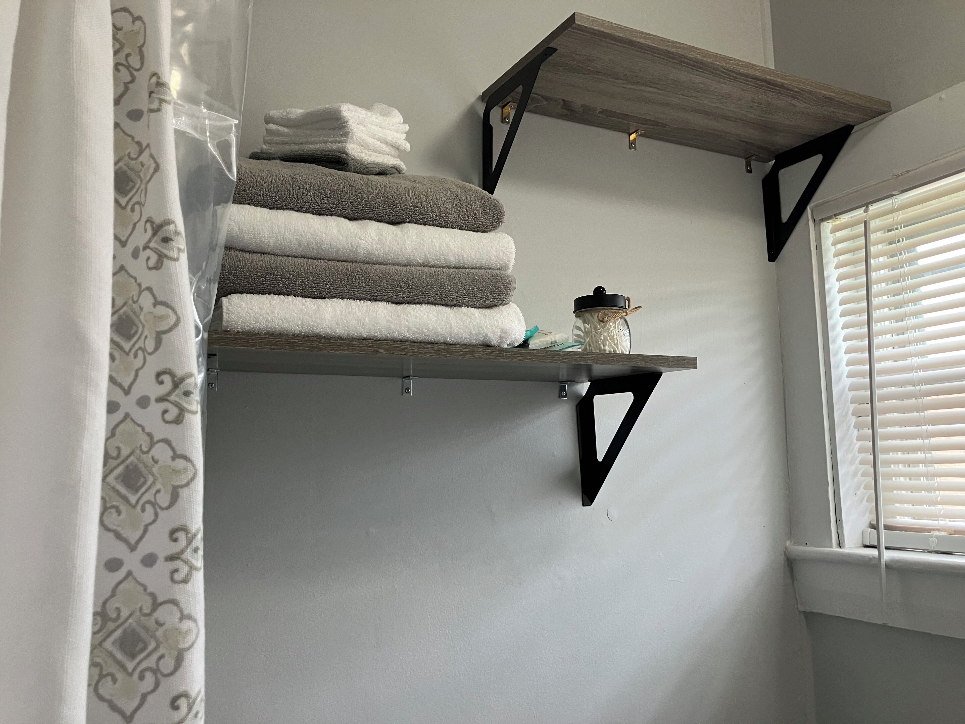A bathroom with a towel rack holding folded towels, a small shelf with a jar, and a window with blinds.
