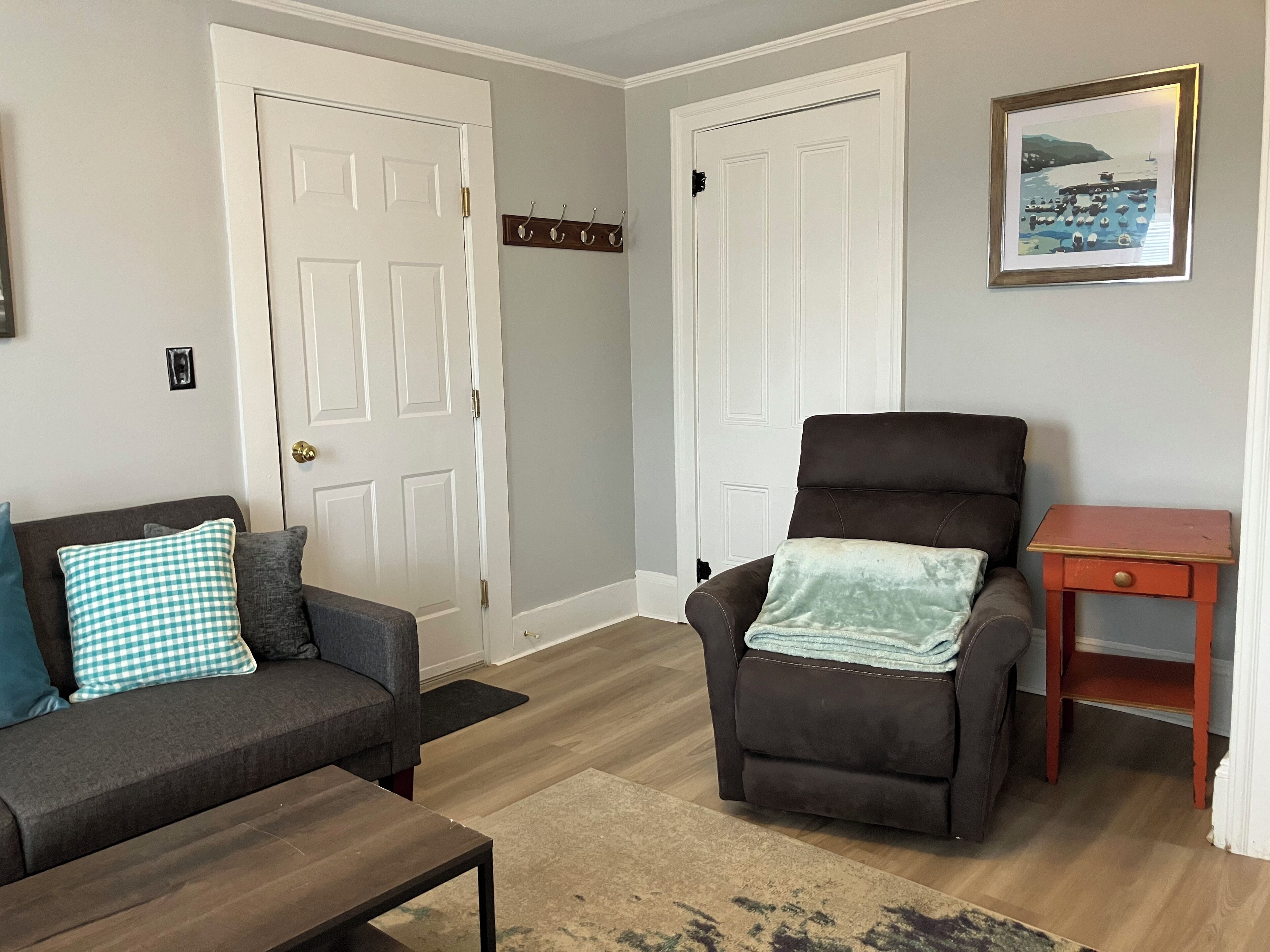 A living room with a dark brown sofa, a light blue and white checkered pillow, a dark brown armchair with a light blue blanket, a small wooden side table, and a framed picture on the wall.