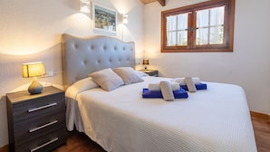 Une chambre à coucher avec un grand lit, une table de chevet avec une lampe, une fenêtre avec des cadres en bois et un tableau accroché au mur.
