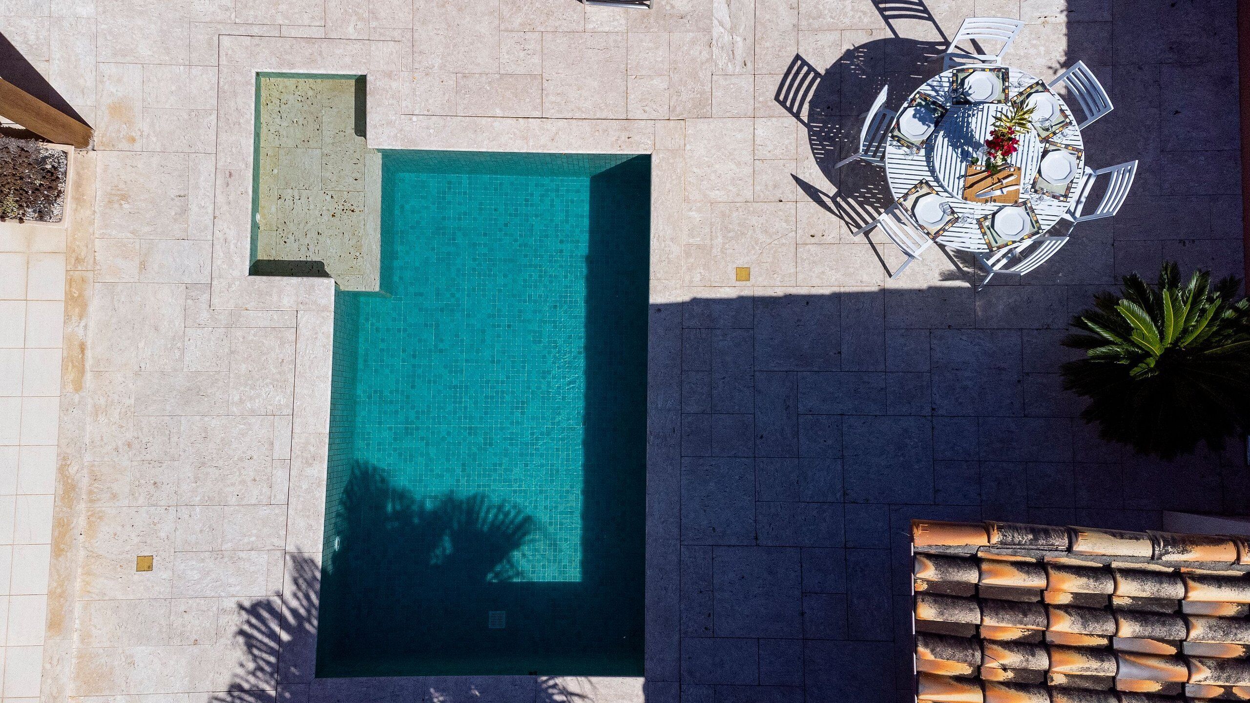 An aerial view of a swimming pool with a tiled deck, surrounded by a patio area with a table and chairs.