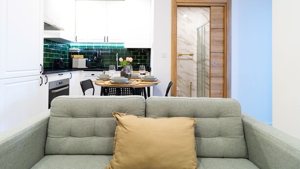 A compact living space with a grey sofa, a small dining table set with plates and glasses, and a kitchen with white cabinets and green backsplash tiles.