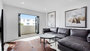 A modern living room with a leather sofa, a glass coffee table, and a large window with a view of buildings.