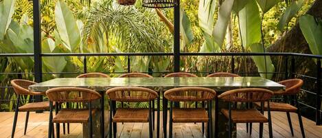 A dining area with a glass table surrounded by wicker chairs, set against a backdrop of lush tropical plants and hanging wicker lamps.