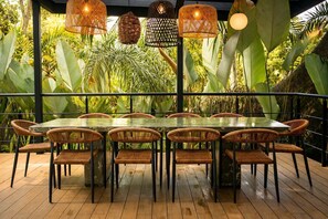 Uma área de refeições com mesa de vidro rodeada por cadeiras de vime, com um pano de fundo de plantas tropicais exuberantes e luminárias de vime penduradas.