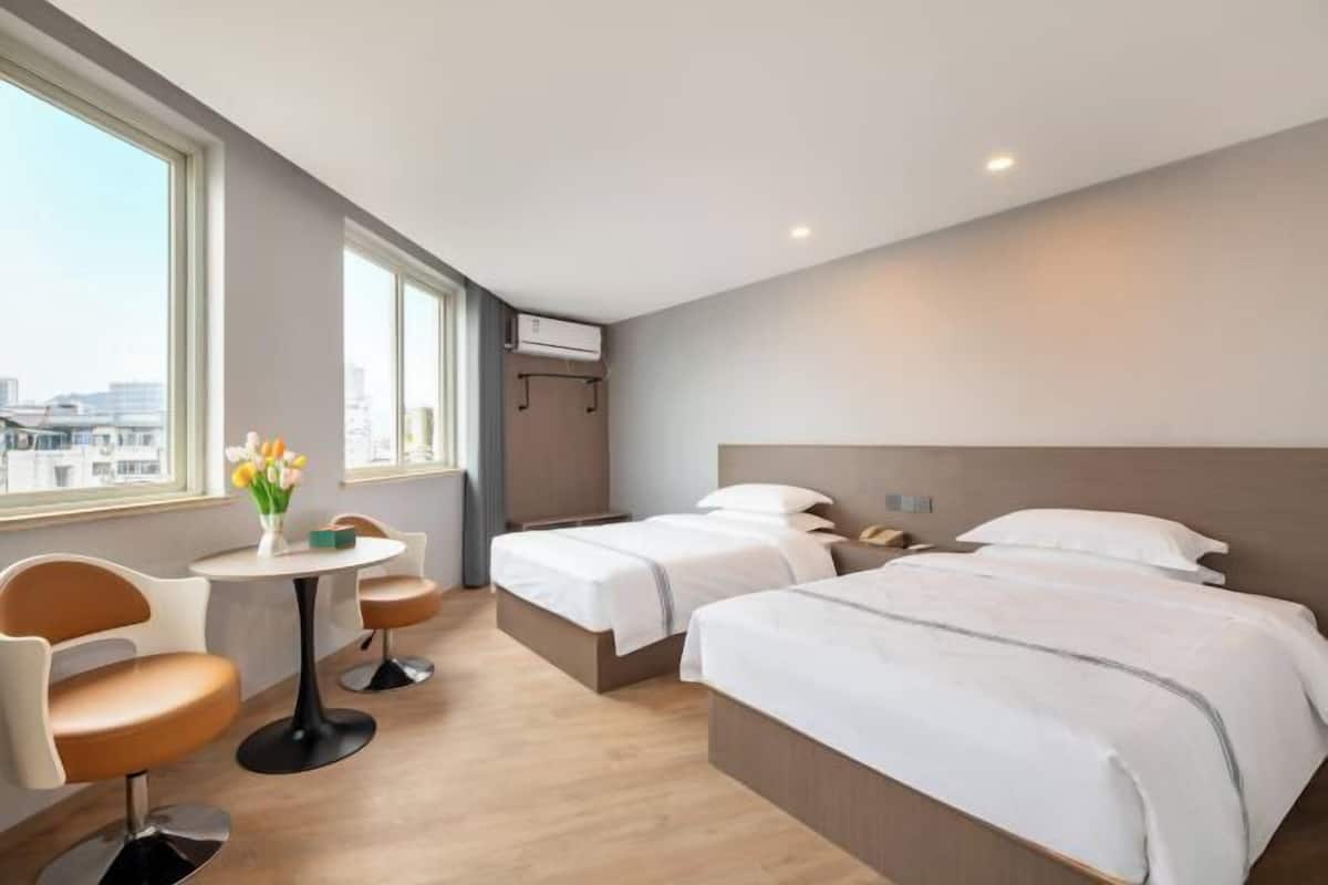 Camera d'albergo con due letti, un tavolo rotondo con sedie, un vaso di fiori e vista sulla città.