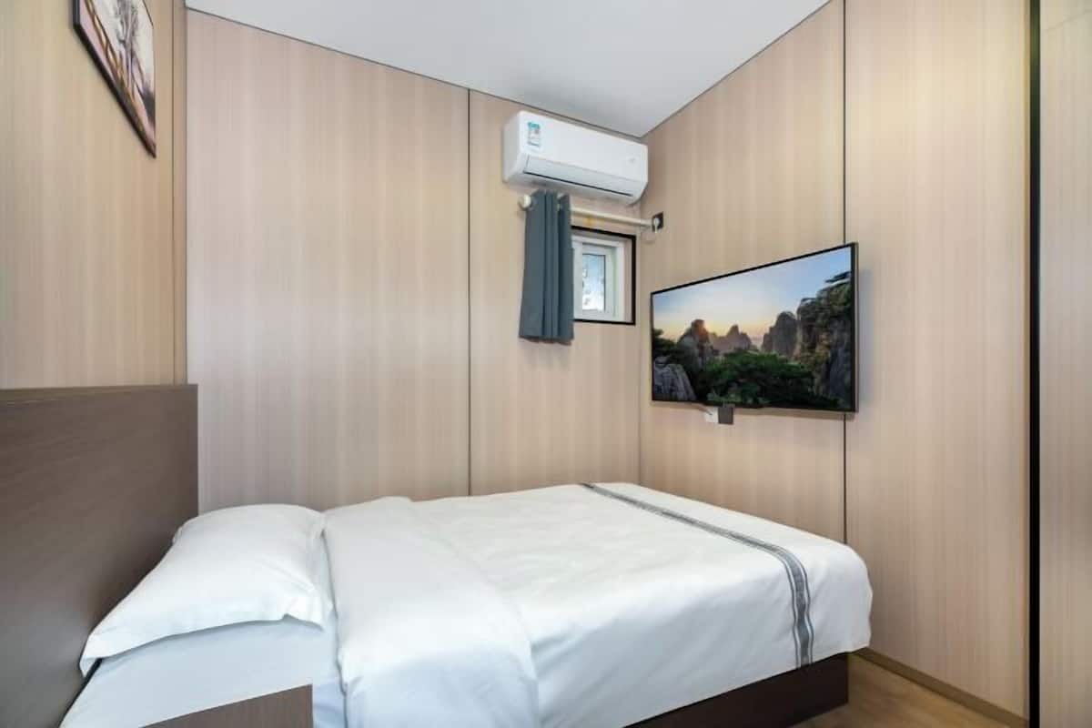 Una camera d'albergo con un letto, un condizionatore d'aria, una finestra con tenda e una TV fissata al muro.