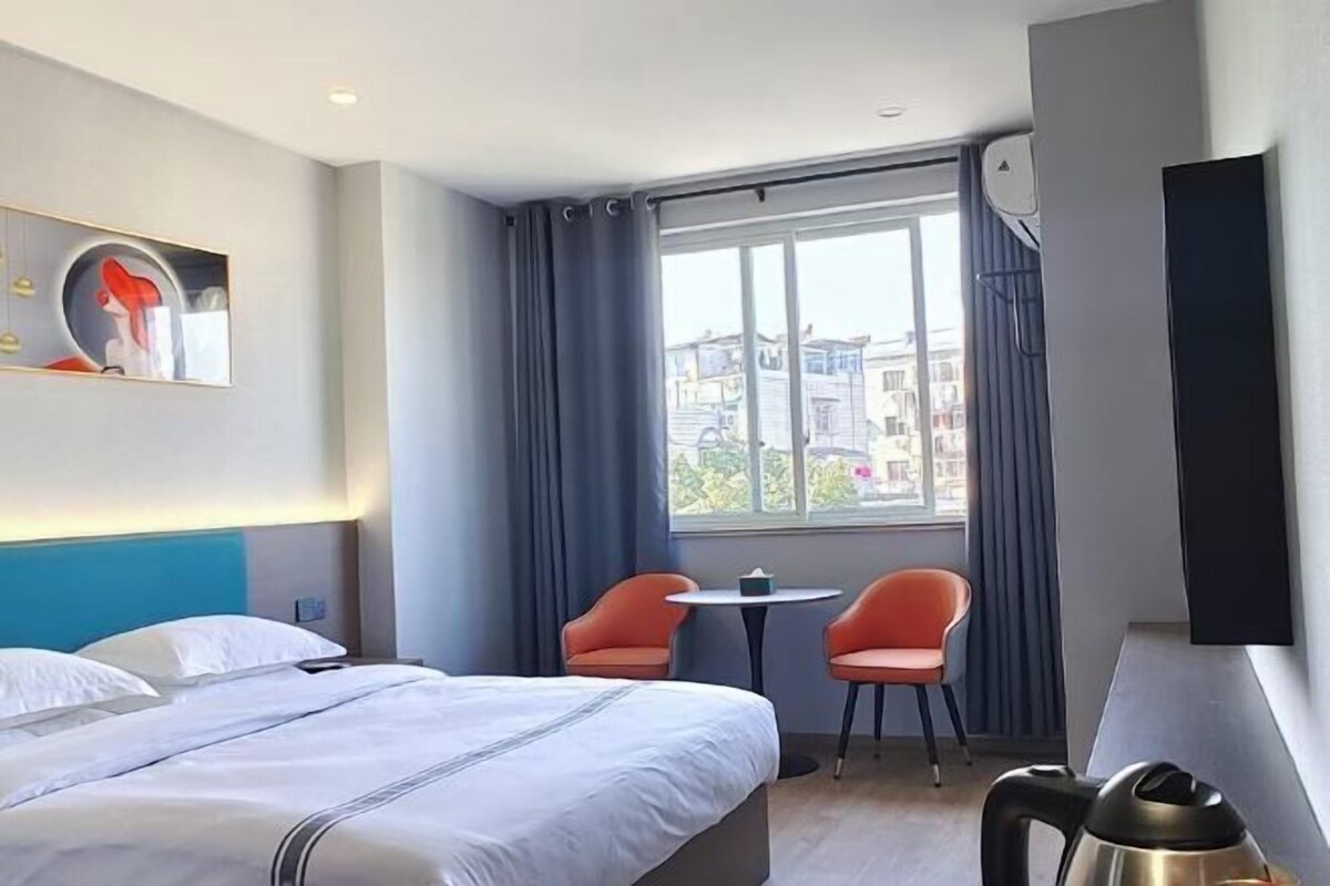 Ein Hotelzimmer mit einem Bett, zwei orangefarbenen Sesseln, einem kleinen Tisch, einem Fenster mit Vorhängen und einem Wasserkocher.