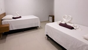 A hotel room with two single beds, each with a white bedspread and a purple towel. There is a wooden headboard on the left bed, and a small nightstand with a lamp. The room has white walls and a tiled floor. - Lençóis Prime Hotel (Barreirinhas)