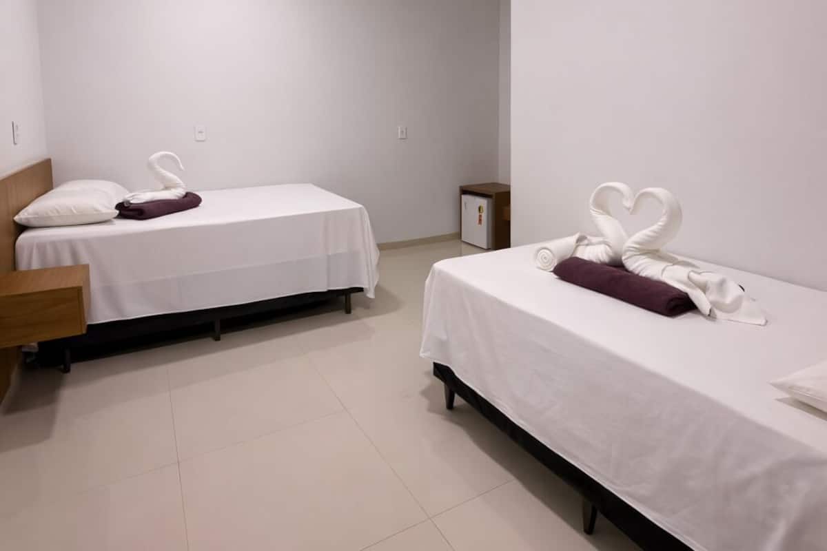 A hotel room with two single beds, each with a white bedspread and a purple towel. There is a wooden headboard on the left bed, and a small nightstand with a lamp. The room has white walls and a tiled floor.