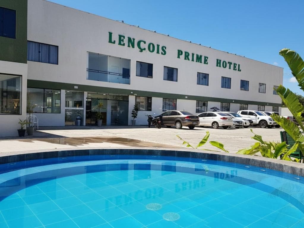 Un hotel con piscina en primer plano, un estacionamiento con varios autos, y un edificio con el nombre 'LENÇOIS PRIME HOTEL' en la fachada.