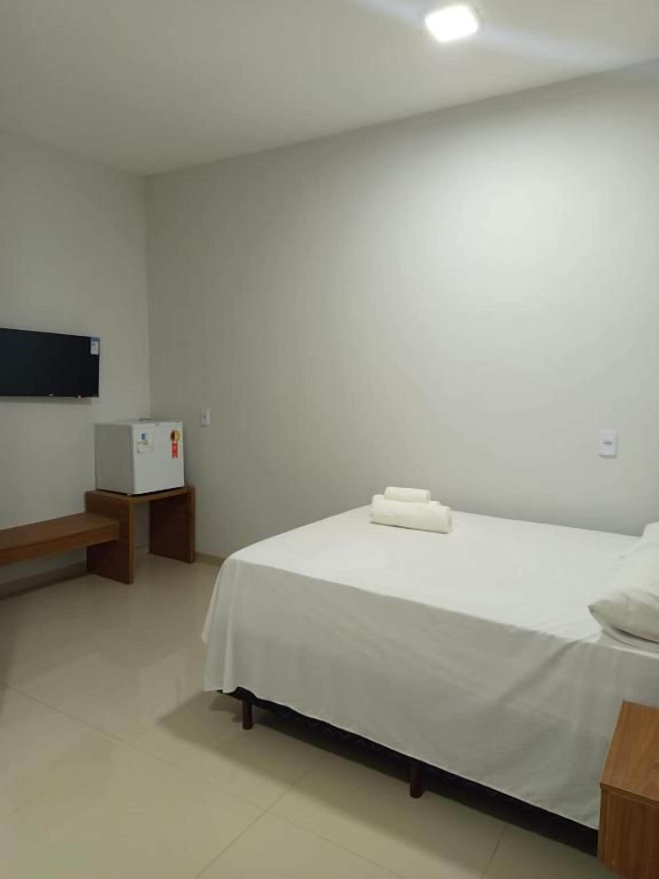 A hotel room with a bed, a small table, a bench, a wall-mounted TV, and a refrigerator.