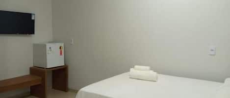 A hotel room with a bed, a small table, a bench, a wall-mounted TV, and a refrigerator.
