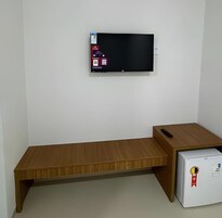 Una habitación con una televisión de pantalla plana montada en la pared, un banco de madera y un refrigerador blanco pequeño.