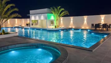 Zwembad met een jacuzzi, omgeven door een witte muur met ingebouwde verlichting, palmbomen en buitengemeubilair.
