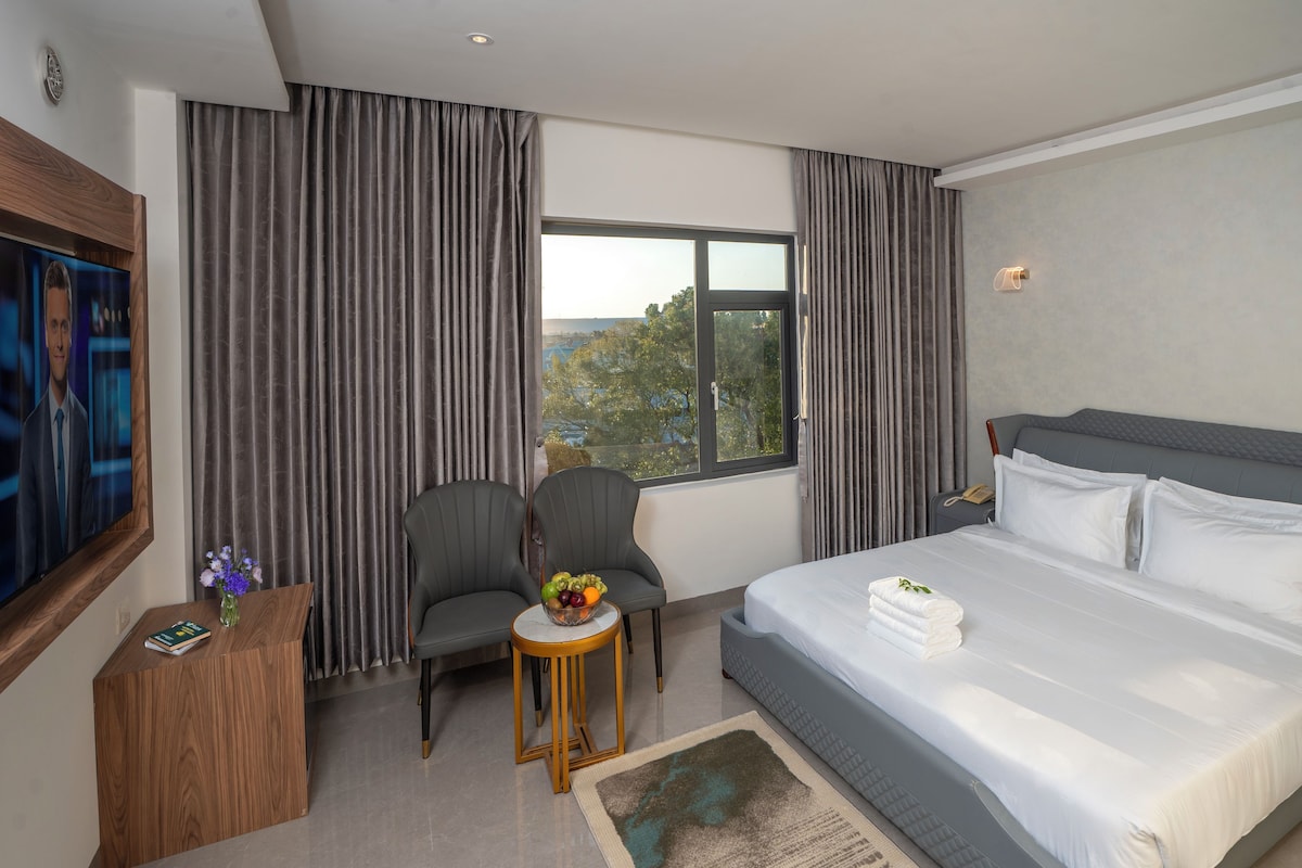 A hotel room with a large bed, two armchairs, a small table with a vase, a painting on the wall, and a window with curtains.