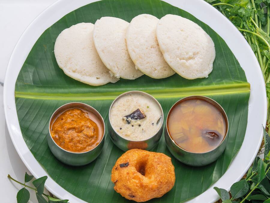 A plate of food with three white idlis, two coconut chutneys, a bowl of sambar, and a fried snack on a banana leaf.
