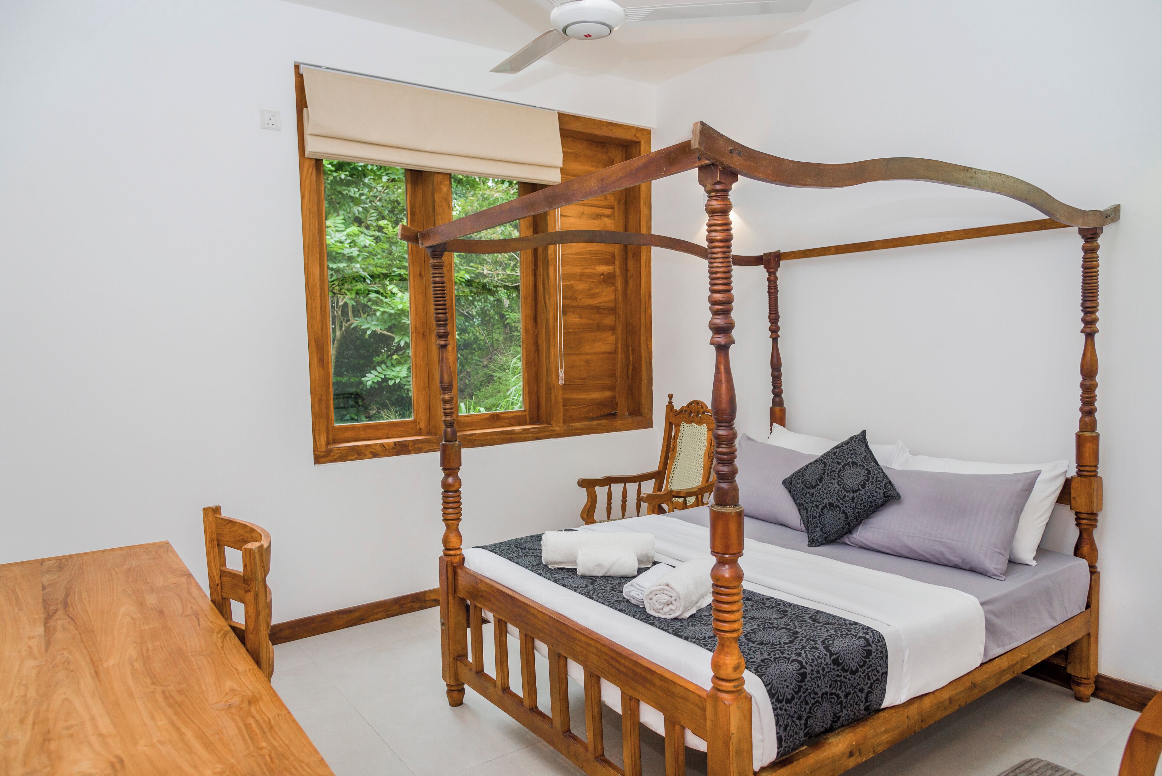 A four-poster bed with a wooden frame, a wooden chair, a wooden table, and a window with a view of greenery.