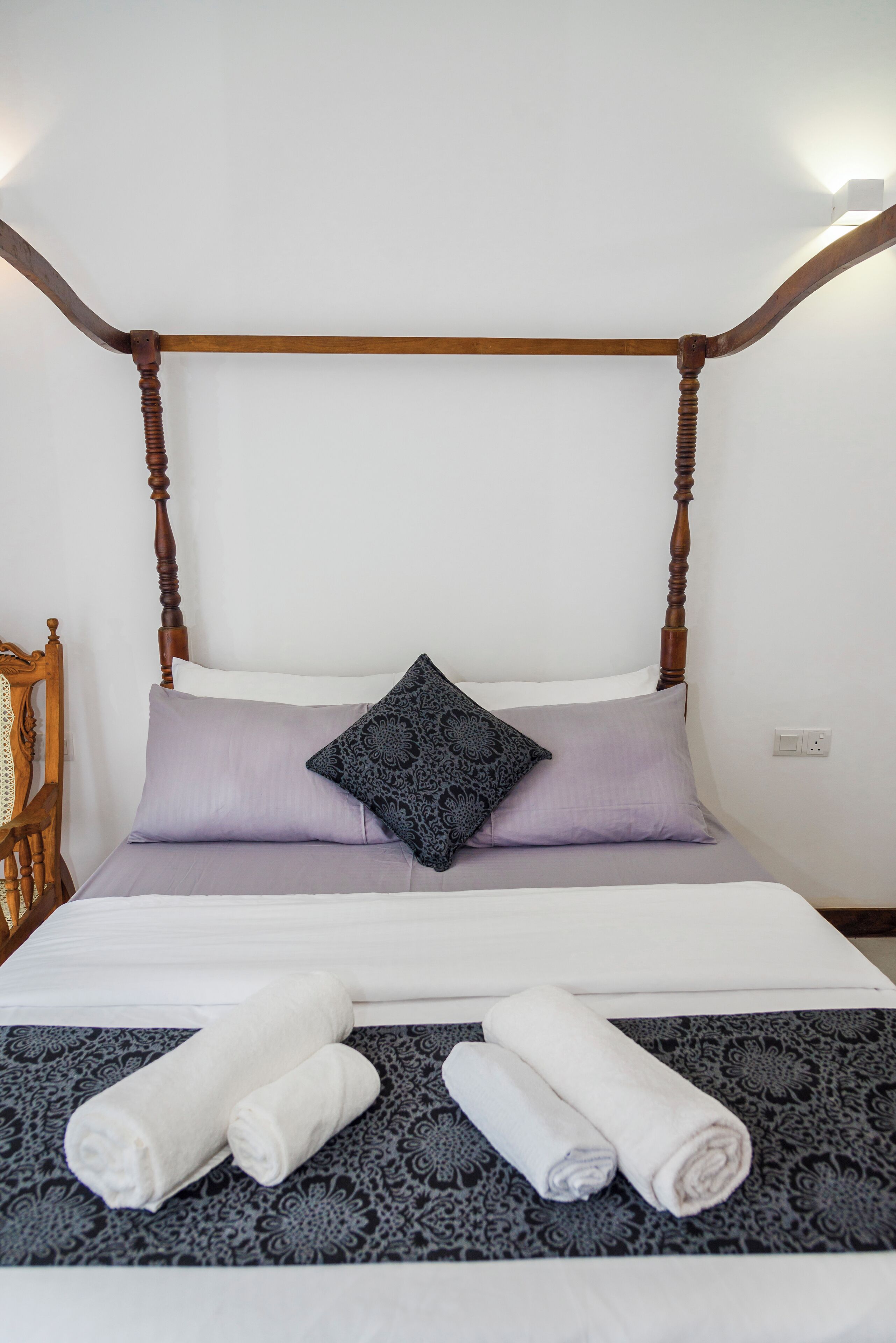 Una cama bien hecha con cabecera de madera, dos almohadas y toallas enrolladas sobre una colcha estampada.