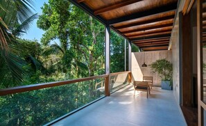 A modern balcony with a wooden ceiling, glass railing, and a view of lush greenery.