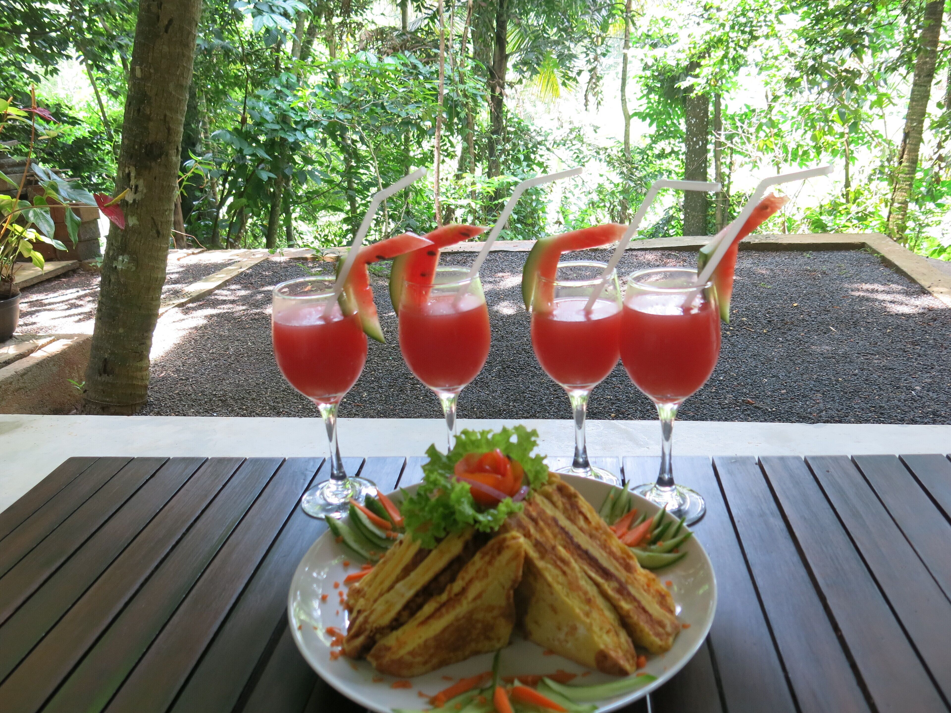A table with four glasses of red drink, a plate of sandwiches, and a view of a forest.
