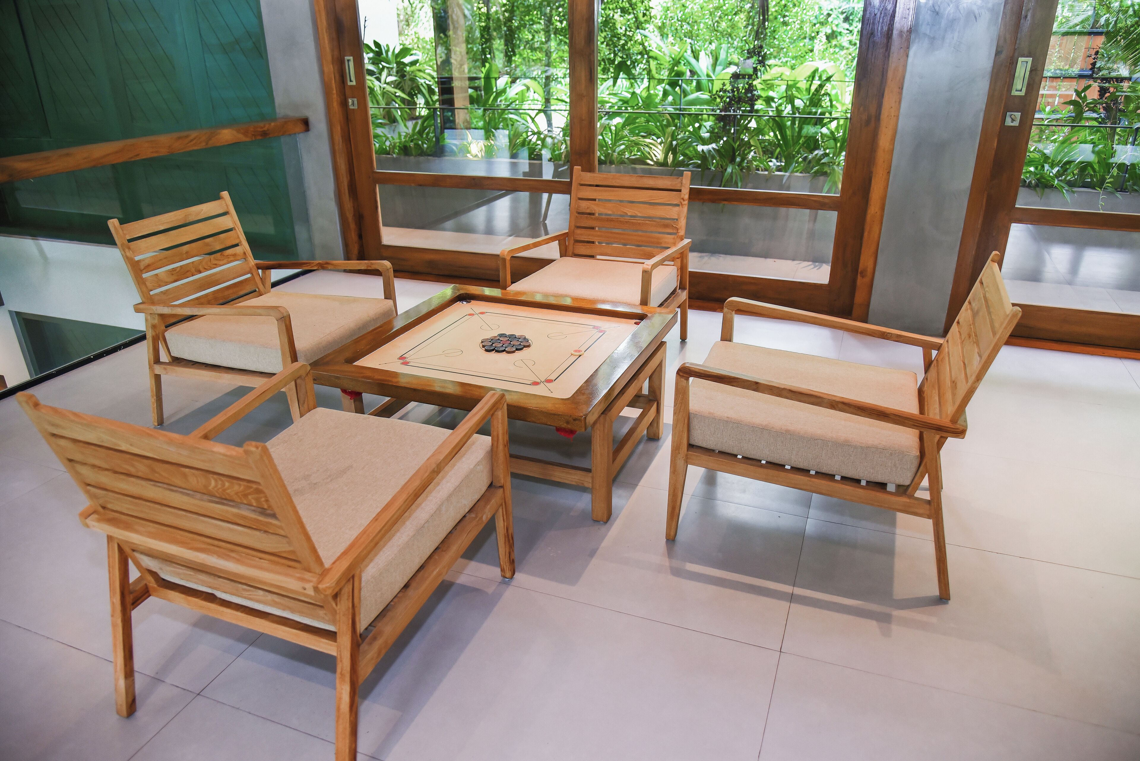 A wooden furniture set with cushioned chairs and a table, arranged in a room with large windows and a view of greenery outside.