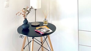 Una sala de estar moderna con un televisor de pared que muestra el logotipo de Netflix, una mesita redonda con una lámpara, un jarrón con flores secas y una revista titulada 'PICCASSO'.