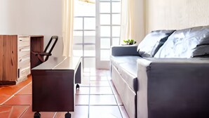 Una sala de estar moderna con un sofá, un escritorio y una silla. Se ve una ventana con cortinas y vistas al exterior.