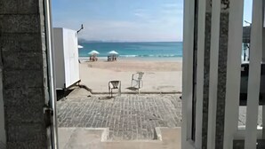 Vista a la playa desde un umbral, con suelo de ladrillo y dos sombrillas blancas.