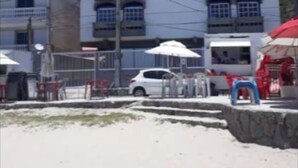 Un edificio de varios pisos con techo plano y balcones, situado cerca de una zona arenosa con mesas al aire libre.