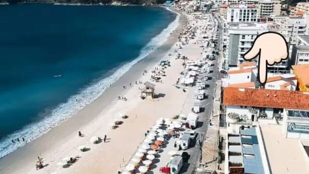 Un paisaje urbano costero con una playa bordeada de sombrillas y tumbonas, edificios con tejados rojos y un telón de fondo montañoso.