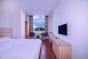 Habitación de hotel con cama, escritorio, televisor, ventana con cortinas y vista a la vegetación.