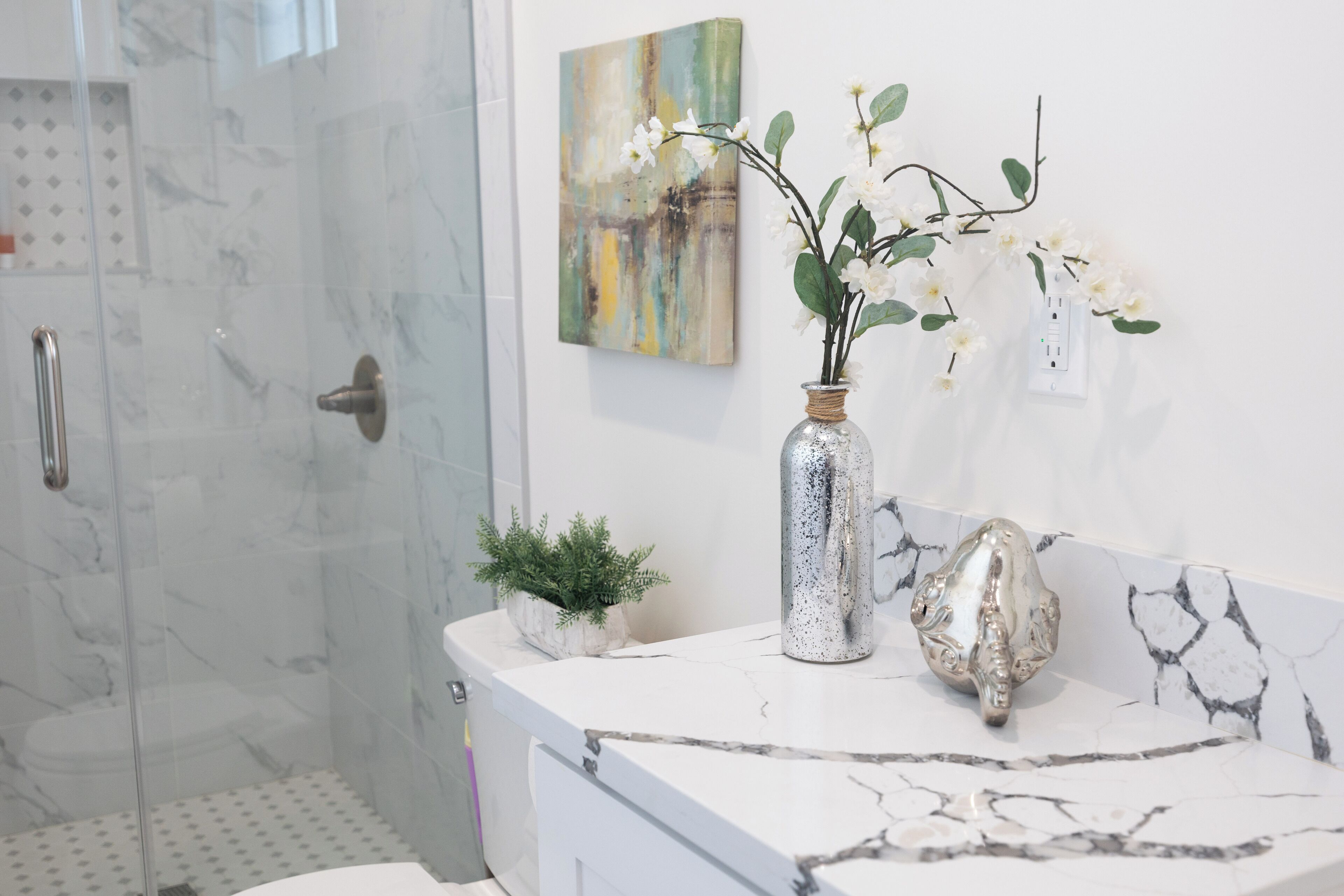 Une salle de bain dotée d’une douche à parois de verre, d’un comptoir en marbre blanc, d’une petite plante en pot, d’un vase décoratif avec des fleurs et d’une prise de courant murale.