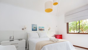 A modern bedroom with a bed, bedside tables, artwork, and a window with a view of trees.
