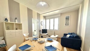 Een eethoek met een ronde houten tafel, witte stoelen en blauwe servetten. Achterin staat een blauwe bank en een kleine witte tafel.