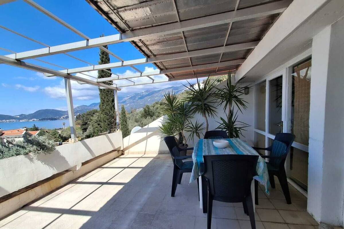 A covered outdoor patio with a table and chairs, overlooking a scenic view of the sea and mountains.