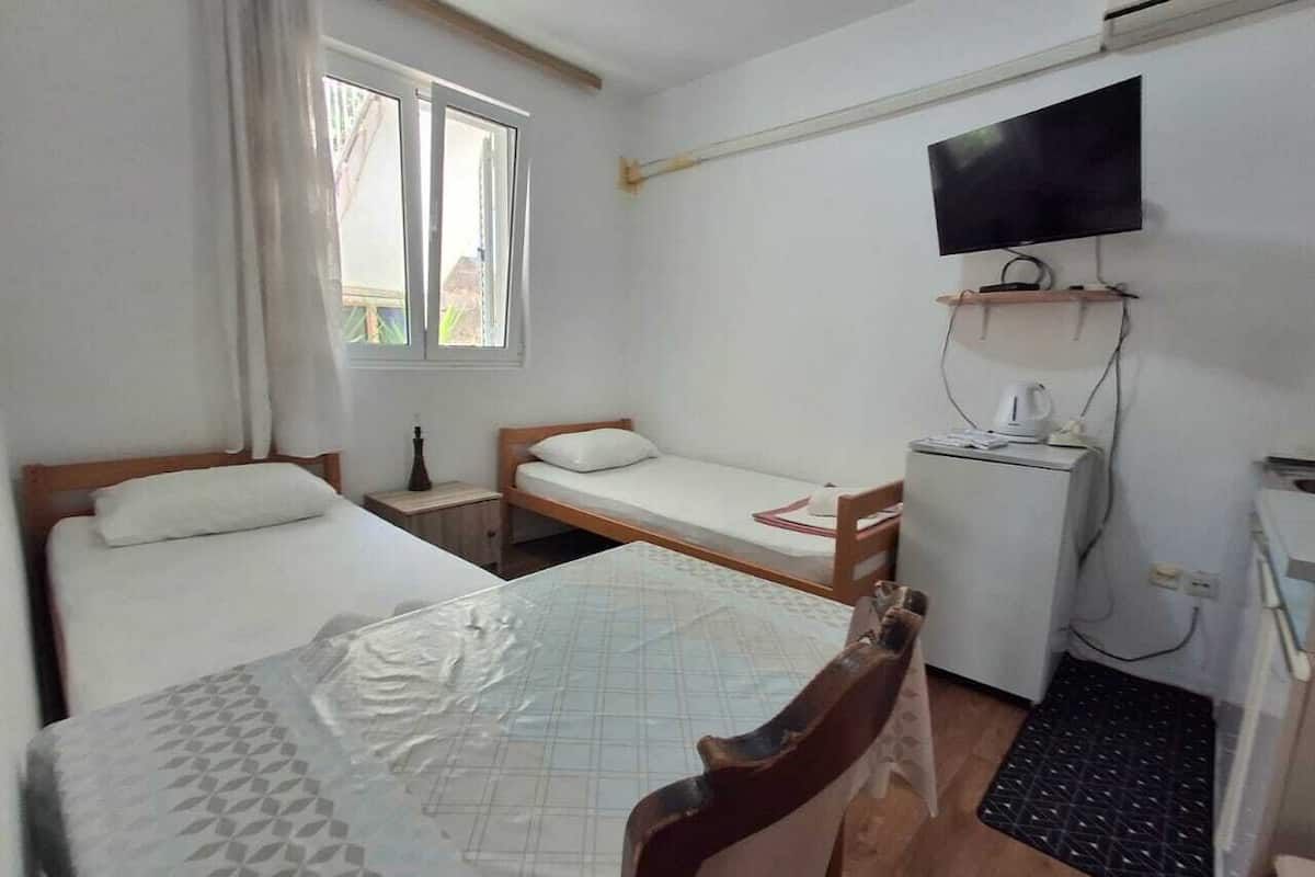 A small, clean room with two single beds, a desk, a chair, a TV mounted on the wall, and a window providing natural light.