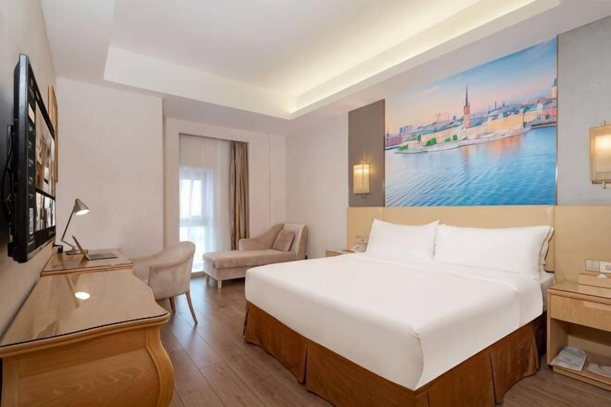 A hotel room with a bed, desk, chair, and a painting of a cityscape.