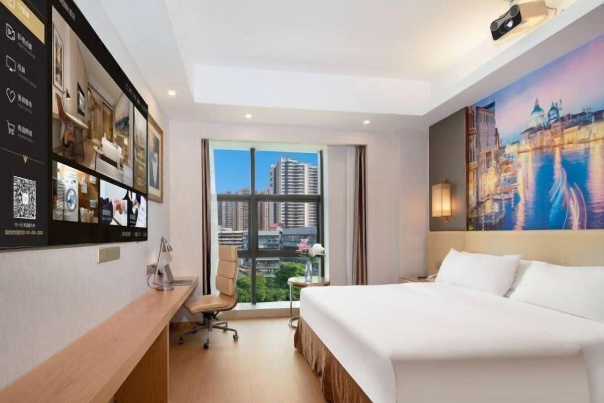 A hotel room with a bed, desk, chair, and a large window with a city view.