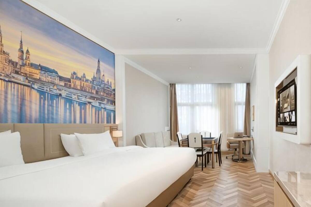 A hotel room with a large bed, a dining table, and chairs, a painting of a cityscape, and a view of a city skyline.
