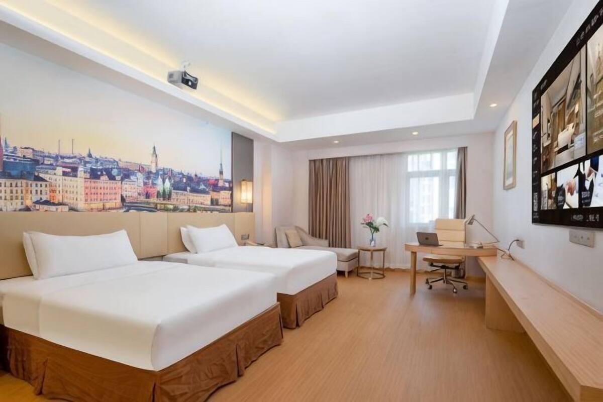 A hotel room with two beds, a desk, a chair, a small table with a vase, and a cityscape mural on the wall.