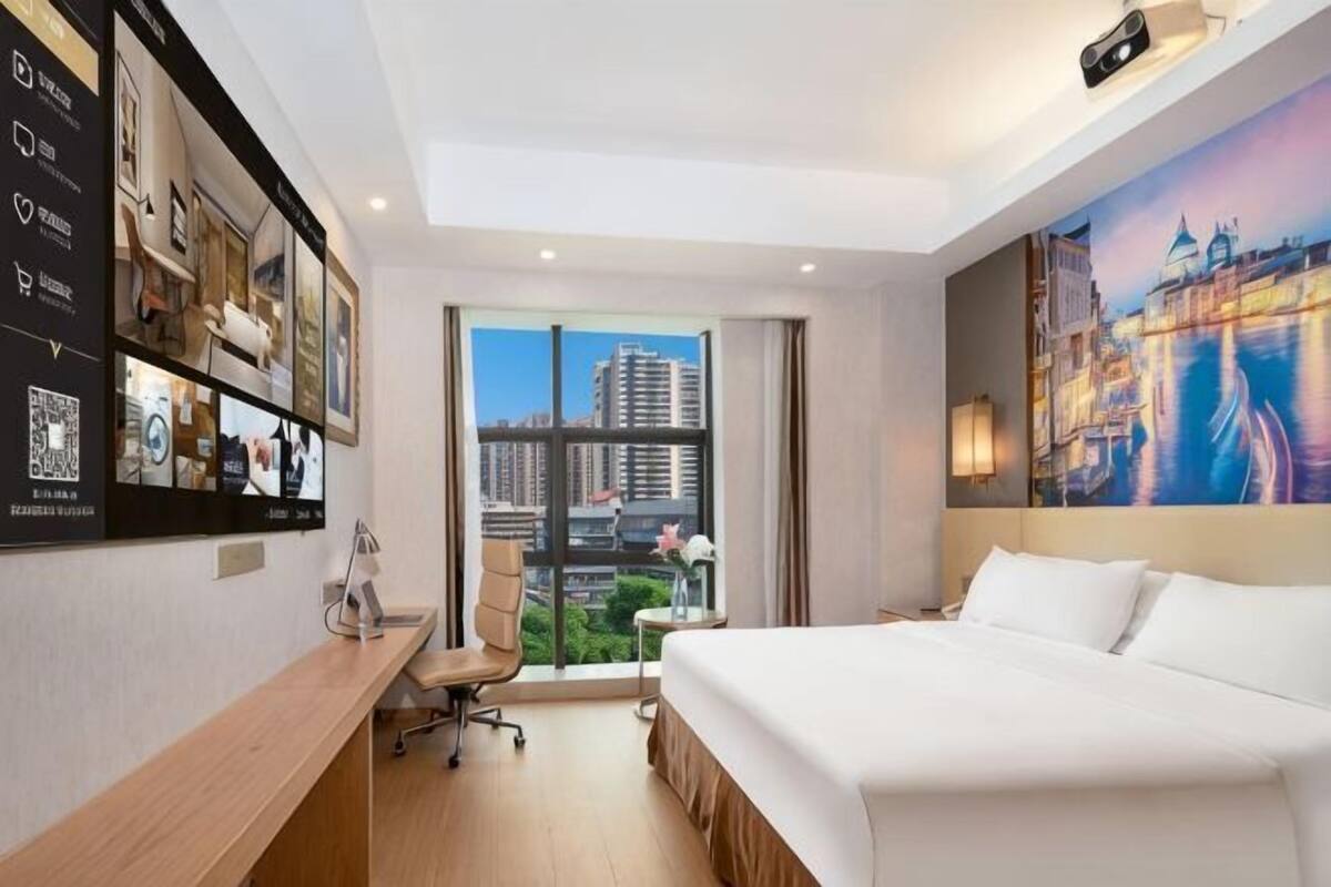 A hotel room with a bed, desk, chair, and a large window with a city view.
