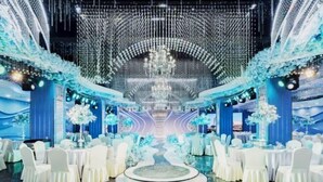 A spacious indoor venue with a central circular platform surrounded by white tables and chairs, and a ceiling adorned with hanging lights and floral arrangements. - Guangming Hotel (Jiuquan)