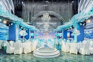 A formal event space with round tables and chairs, a central decorative feature, and a backdrop with a scenic view. - Guangming Hotel (Jiuquan)