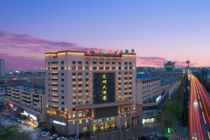 A multi-story hotel with a prominent sign, surrounded by other buildings and a busy road with light trails. - Guangming Hotel (Jiuquan)