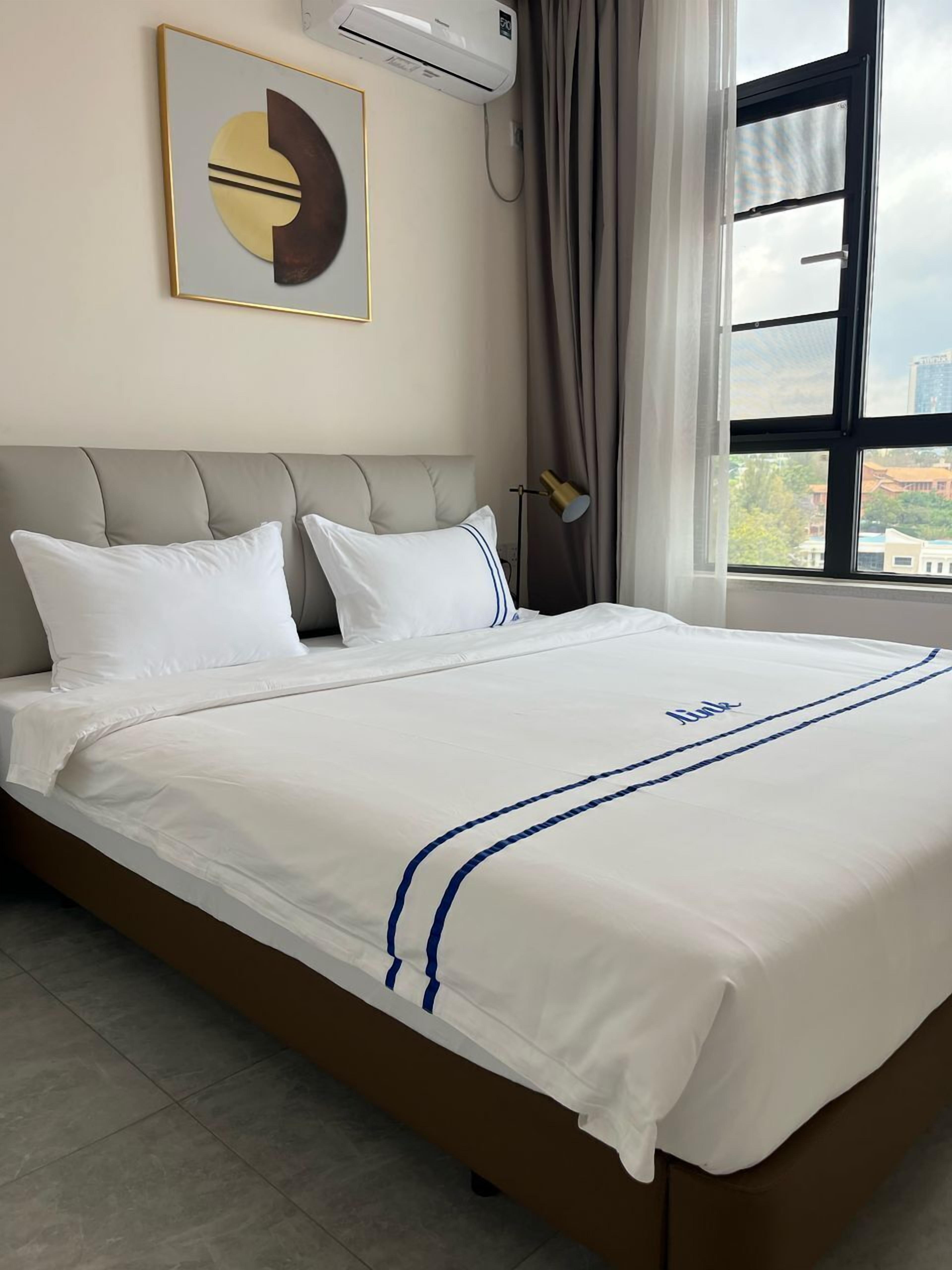 Ein ordentlich bezogenes Bett mit weißer Bettwäsche und blauen Streifen, ein moderner Kopfteil und ein großes Fenster mit Blick auf die Stadt.