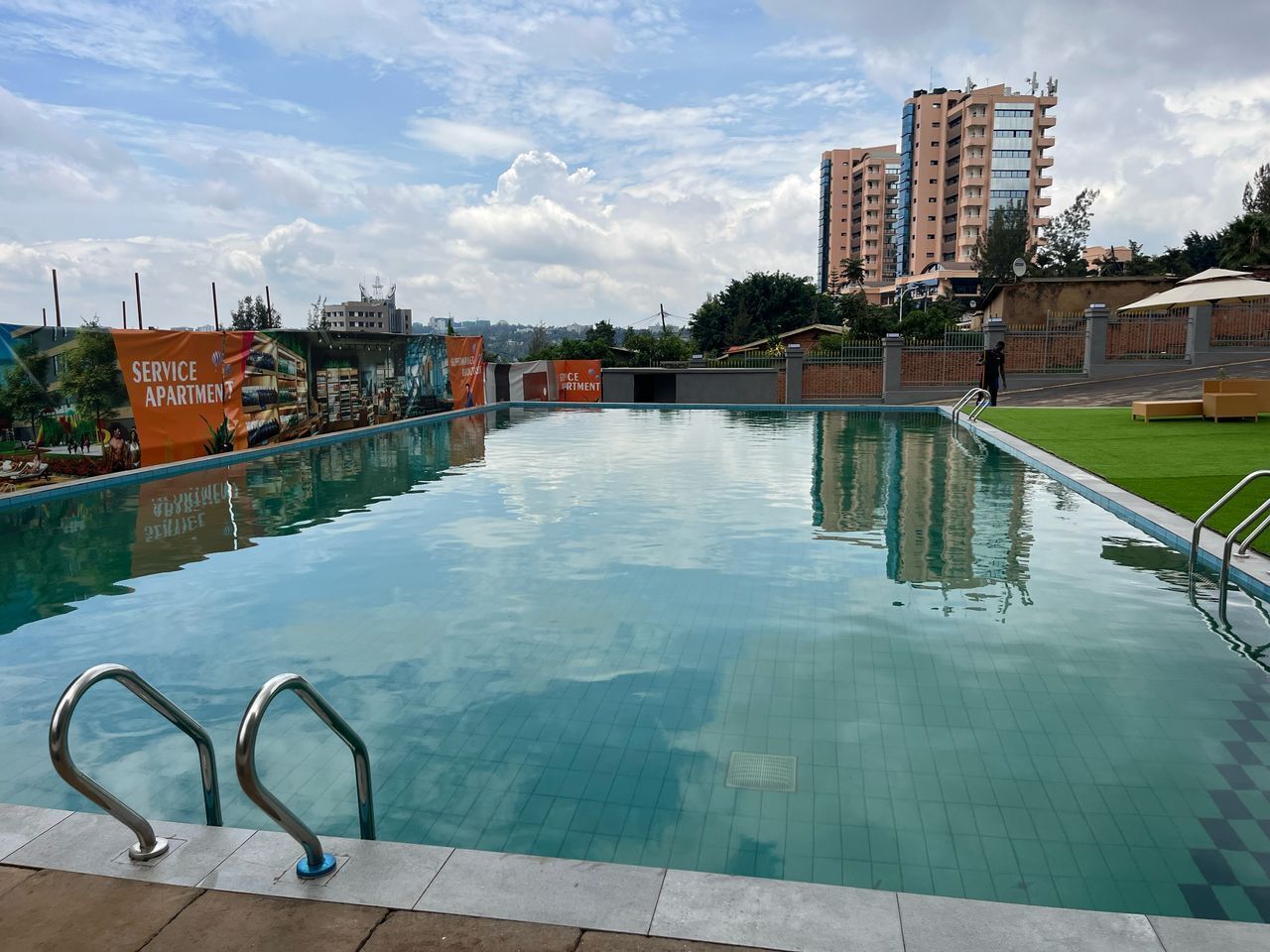 A swimming pool with clear water, surrounded by a tiled deck and metal handrails. In the background, there are buildings and a grassy area.