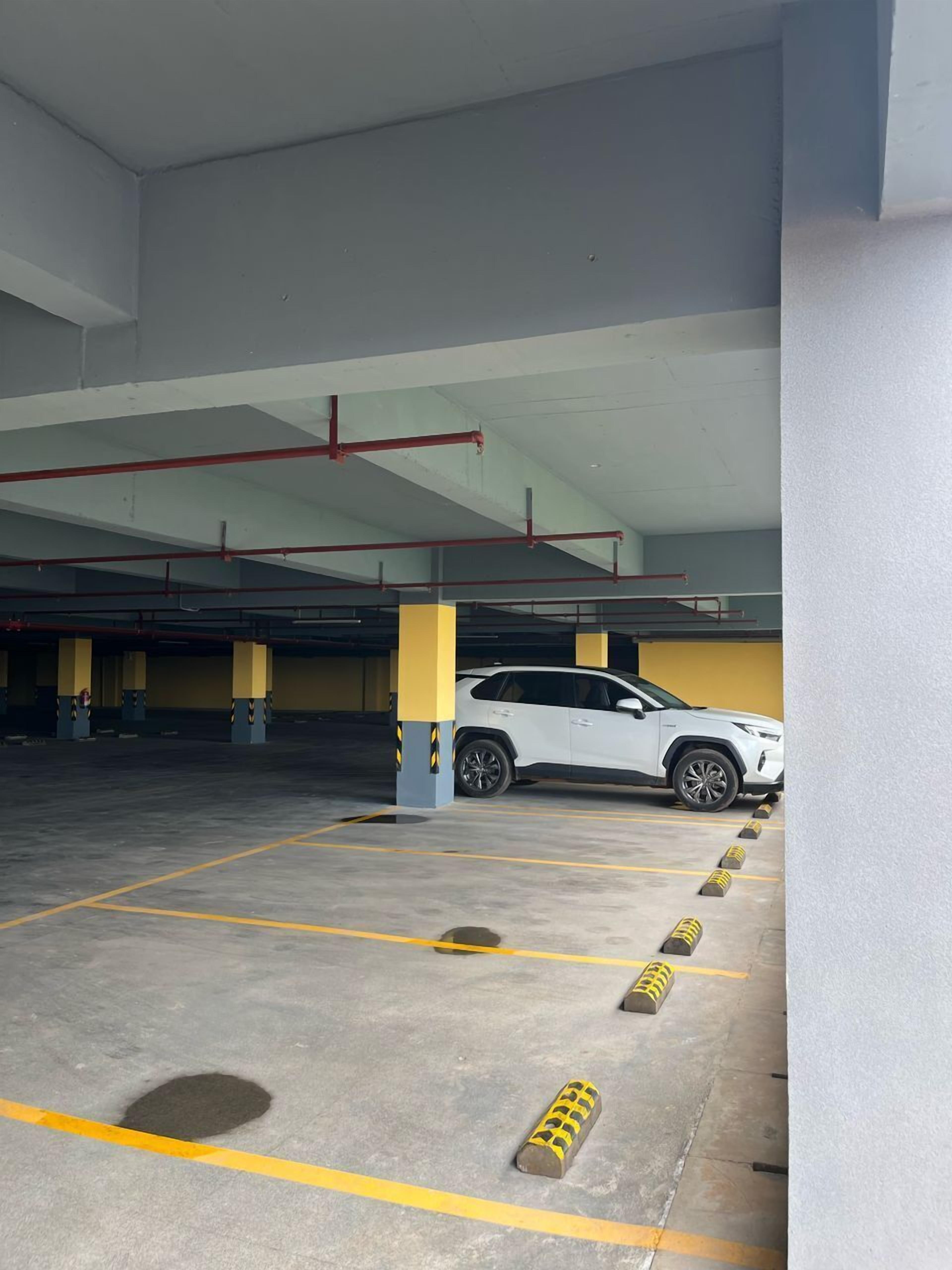 Un aparcamiento de varios niveles con pilares amarillos y grises, una camioneta SUV blanca estacionada en un hueco, y bolardos amarillos y negros a lo largo de las líneas de estacionamiento.