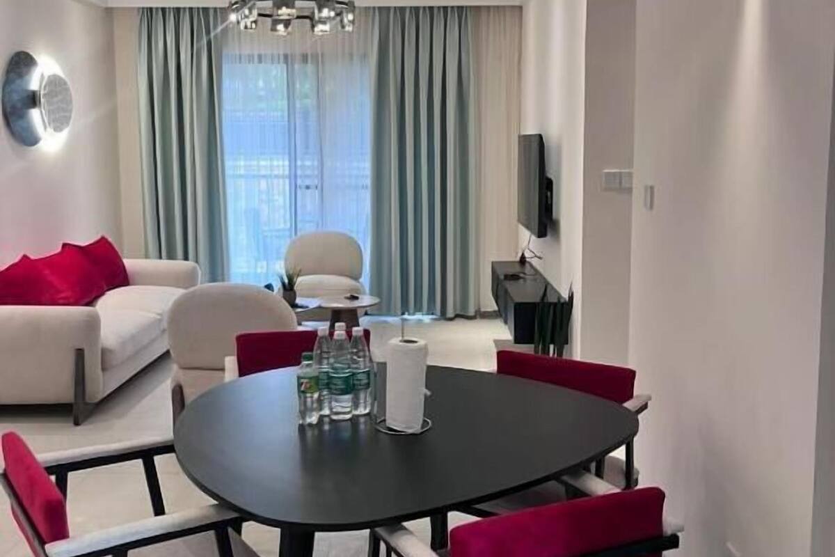 Een moderne woonkamer met een ronde eettafel, rode en witte stoelen, een bank, een televisie en een kroonluchter.
